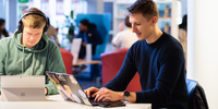 Two students working at their laptops at a shared desk in the library