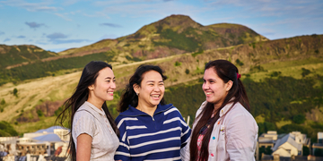 Three students standing at the bottom of Arthur's Seat on a sunny day, laughing with each other