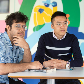 Two students sitting in the common room with books in front of them, listening to someone talk