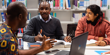 Three students discussing work over laptops and notes in a study area