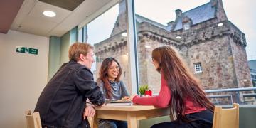 Three students chatting at a table in a bright room next to Merchiston Tower