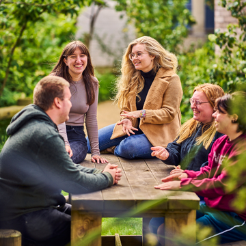 A group of students sitting at a picnic table outside, with green trees in the background