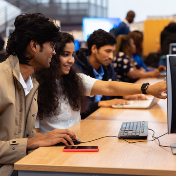 A group of students sitting at a row of computers, pointing at something on the screen