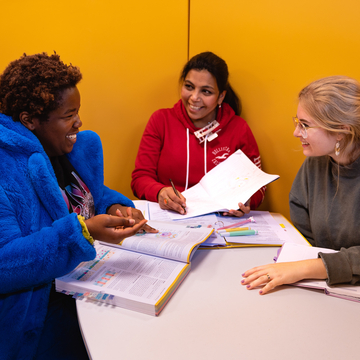Three students smiling and talking in a yellow study pod, with books and notes in front of them