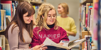 Two students looking over an open book between shelves at the library