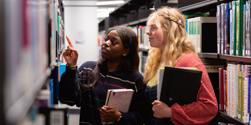 Two students holding books and looking at bookshelves in library