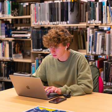 Student at a desk in the library in front of some bookshelves, studying on a laptop