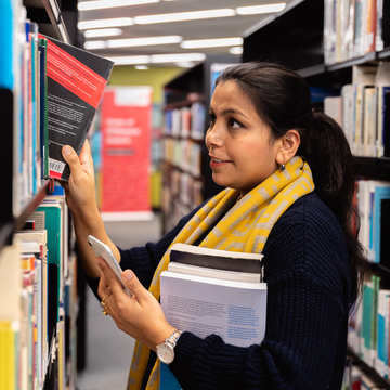Student holding a pile of books and taking a book from the library shelves
