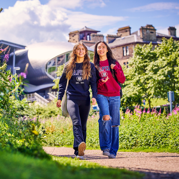 Two students wearing Edinburgh Napier jumpers walking through Craiglockhart grounds in the sun, with wildflowers in the foreground and the lecture theatre in the background