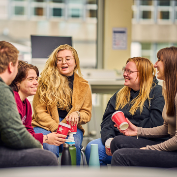 Group of five students sitting chatting in campus cafe, holding coffee cups