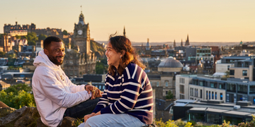 Two students sitting in front of a view of Princes Street and Edinburgh Castle at sunset