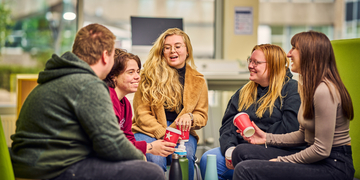A group of students sitting talking to each other in a cafe on campus, some holding red coffee cups