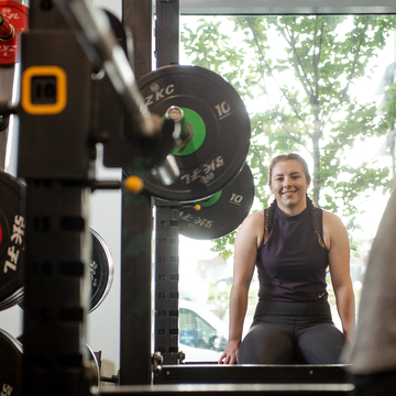 Student in gym clothes sitting next to a bright window, beside olympic weighlifting rack