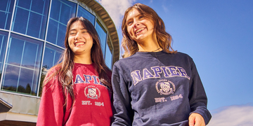 Two students outside campus on a sunny day, one wearing a maroon Edinburgh Napier hoodie and one wearing a navy version
