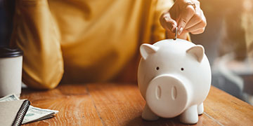Person in a yellow jumper sitting at a table, depositing a coin into a white piggy bank