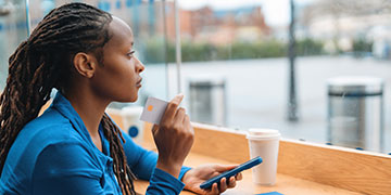 Student sitting at a high table with a coffee, holding a mobile phone and debit card