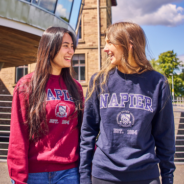 Two students standing outside Craiglockhart campus in the sun, wearing maroon and navy Edinburgh Napier hoodies