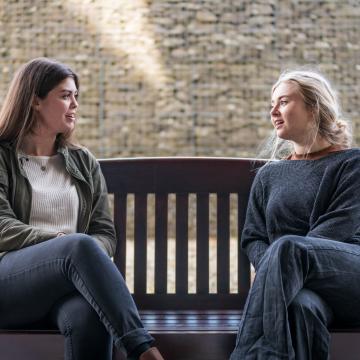 Two students talking to each other on a bench next to a wall outside