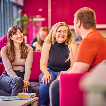 Three students sitting on pink sofas, laughing and chatting to each other