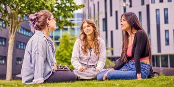 Three students sitting on the grass talking outside campus, with the building in the background
