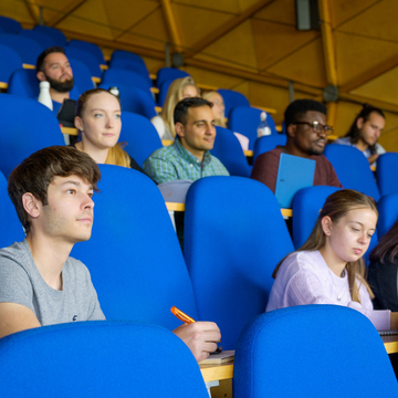 Students sitting on blue chairs in a lecture theatre as they listen and write notes