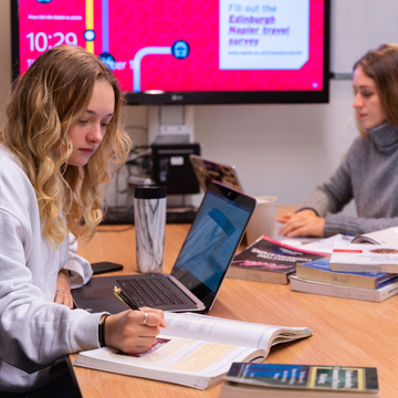 Two students sitting in a study space, working together with laptops and notes
