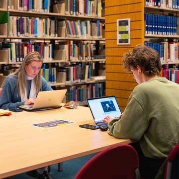 Two students sitting in the library, working opposite each other with laptops