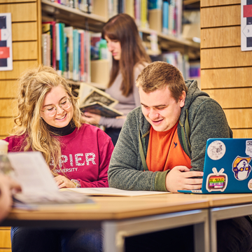 Two students sitting in a study space, working together with laptops and notes