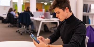 Student sitting at a desk in the library, looking at the Edinburgh Napier app on a mobile phone