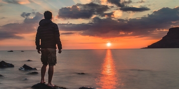 Student in hoodie and shorts standing barefoot on a rock at the sea, looking out over the water while the sun sets