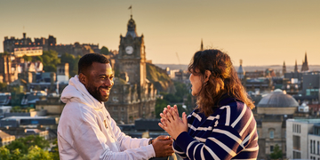 Two students sitting and laughing in front of a view of Princes Street and Edinburgh Castle at sunset