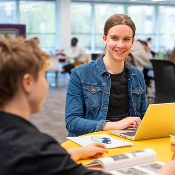 Two students sitting at a table in the library, talking and working with books and laptops
