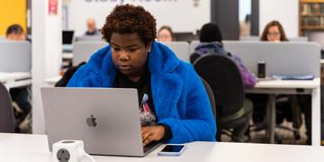 Student in a bright blue coat working at a laptop in a computer suite