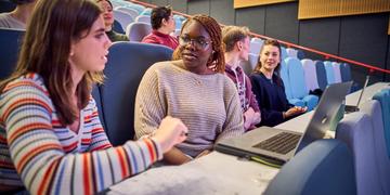Students sitting in a row in a lecture theatre, looking at notes and a laptop