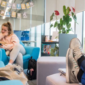 Image of female sitting on comfortable seating in the relaxation space in Sighthill Library. In the foreground, there is a close up of a pair of trainers as someone rests their feet on a stool