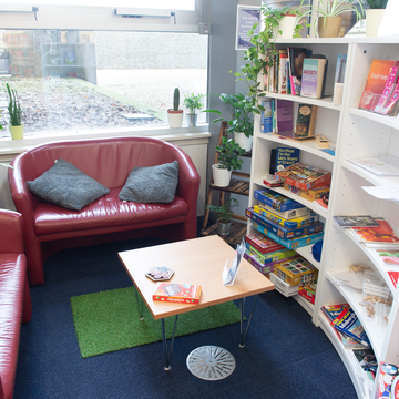 Craiglockhart Library relaxation space. Two small sofas to the left and back of the image in front of a window. A  a curved set of white bookshelves are to the right of the image. On the bookshelves are games and puzzles and books. A low table is in the centre of the image.