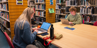 Two students sitting at a desk in the library, both working on their laptops