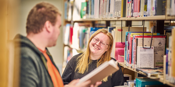Two students standing in between library shelves, talking to each other while holding books