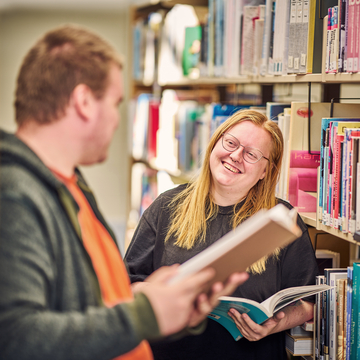 Two students standing between library shelves, holding books and talking to each other
