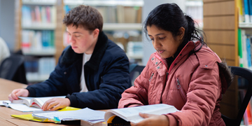 Two students sitting next to each other at a library desk, looking at their books
