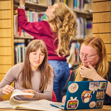 Two students working with a laptop and notes at a desk in the library, with another student looking at books on the bookshelf in the background