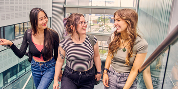 Three students talking to each other as they walk up a stairway on campus, with a bright foyer in the background