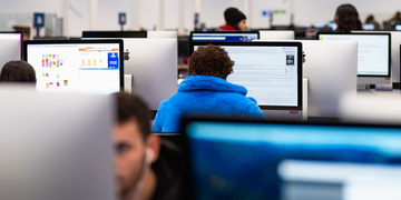 Student in a blue hoodie sitting at a screen in the computer suite