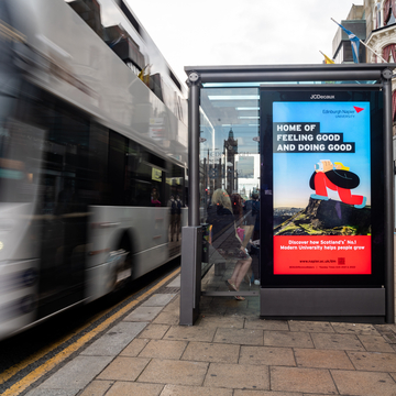 Bus stop with Edinburgh Napier poster in window. A bus is pulling alongside the stop, slightly out of focus.