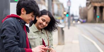Two students standing on Princes Street next to tram tracks and bus lane, looking at a phone together