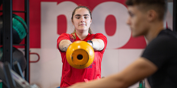 Student in a red t-shirt in gym, lifting an orange kettlebell with both hands