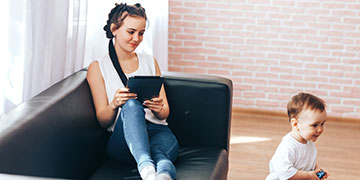 Student sitting on the sofa with a tablet in hand, watching a toddler play next to her