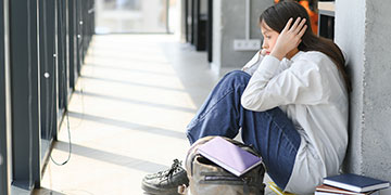 Student sitting in a corridor with her hands over her head, with her bag and books around her