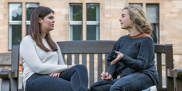 Two students sitting on a bench outside campus, talking to each other