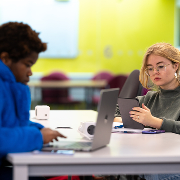 Two students sitting in a study space, working together with laptops and notes